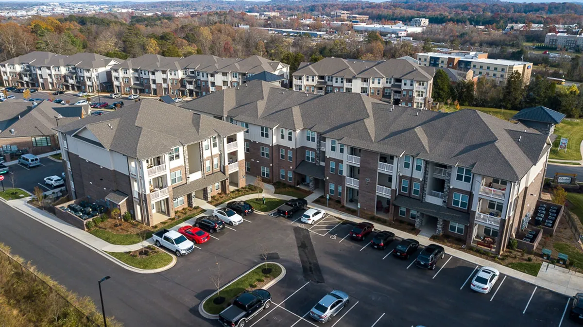 This image shows modern apartment buildings surrounded by a well-maintained parking lot and lush green spaces. The exterior is friendly and welcoming, with areas where children can play and families can gather. The landscaping and clear walkways suggest a safe, community-oriented environment ideal for families.