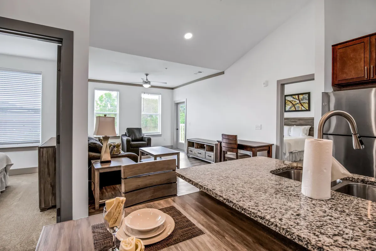 This image showcases a spacious open-plan area that combines a living area with comfortable seating and a dining set adjacent to a modern kitchen featuring a granite countertop. Large windows fill the space with natural light, and the overall look is contemporary and welcoming, with carefully chosen decor.