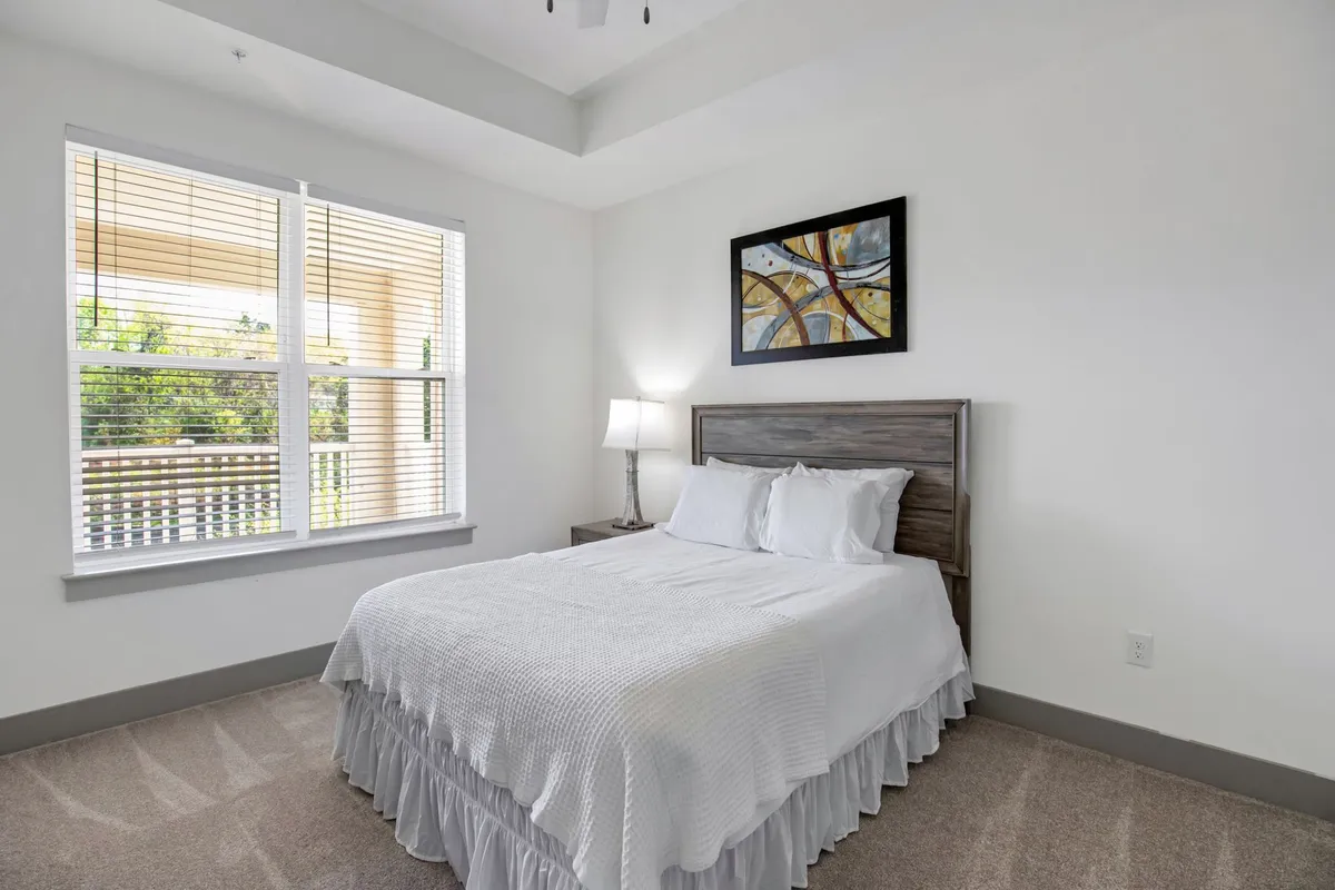 A serene bedroom with a comfortable bed, soft lighting from a bedside lamp, a large window allowing light to filter in, and tasteful wall art. The space is organized and calming, ideal for restful nights or quiet reading.