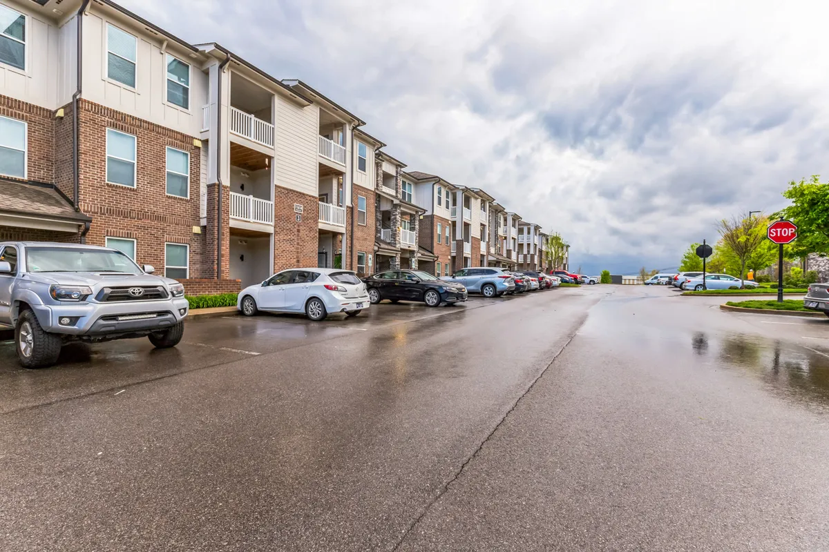 A tranquil street scene featuring apartment buildings, parked cars, and a visible street sign. The area looks pedestrian-friendly, with people walking and a sense of neighborhood life. The surrounding environment suggests proximity to urban amenities and a pleasant, walkable setting.