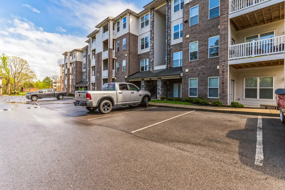 A modern apartment building is featured with a paved parking lot in the foreground, bordered by green landscaping. A few vehicles are parked, and people are seen engaging in casual social activities around the outdoor spaces, including the courtyard. The scene is welcoming and suggests a vibrant but relaxed community atmosphere.