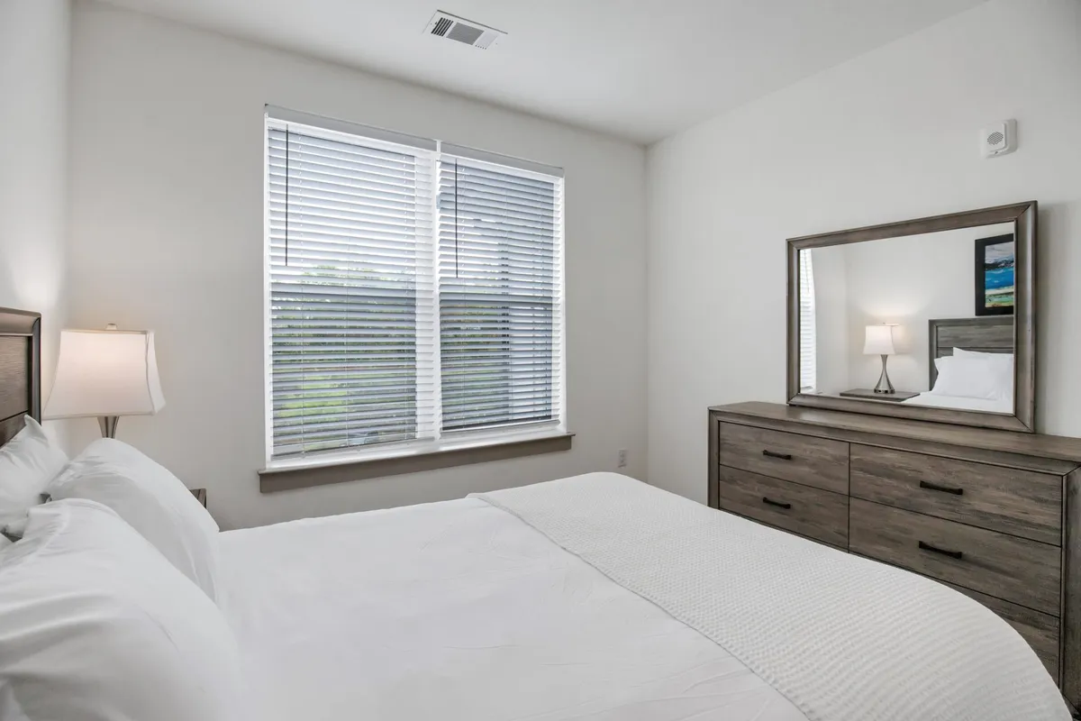 An interior view of a tranquil bedroom with a comfortable bed, a window allowing in natural light, and a mirror adding depth. The space is designed for rest, reading, or relaxation, evoking a sense of calm and comfort at the end of a busy day.