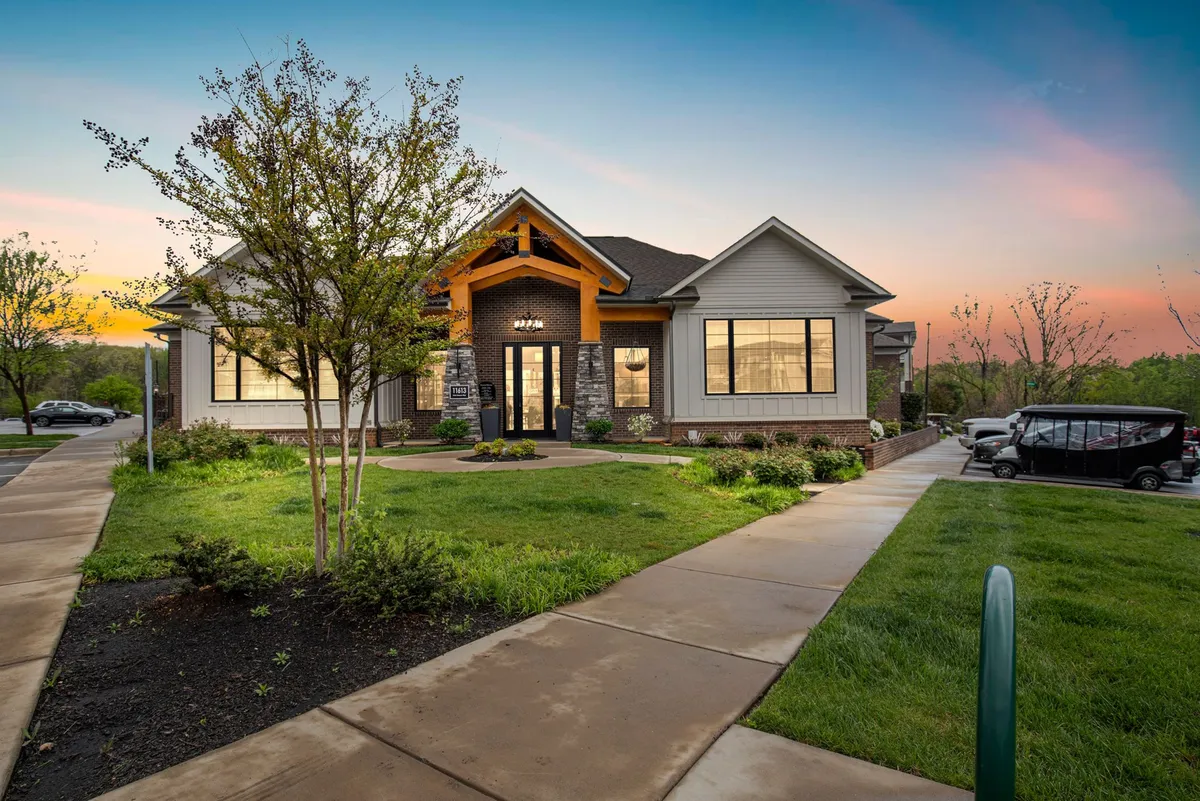 The image showcases the modern exterior of the Overlook at Farragut apartment, with striking architectural lines, expansive windows, and a blend of stone and wood accents. Lush, landscaped greenery surrounds the building, and a sunset glow adds warmth. The scene hints at community spaces perfect for gatherings, relaxing evenings, and scenic strolls.