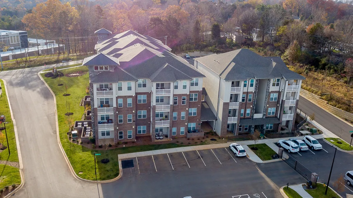 A view of modern apartment buildings with contemporary architecture, landscaped grounds, and a parking area. The image showcases the inviting entryway, lush greenery, and the overall welcoming atmosphere of the community.