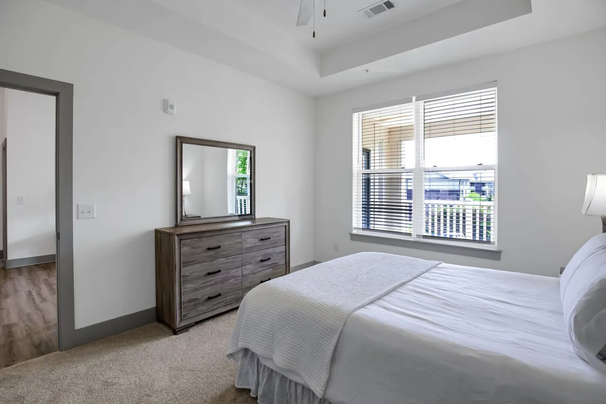 A second bedroom is highlighted here, with a neatly made bed, a dresser topped with a mirror, and large windows that allow natural light to bathe the space. The décor is simple yet elegant, reinforcing a restful, inviting ambiance.