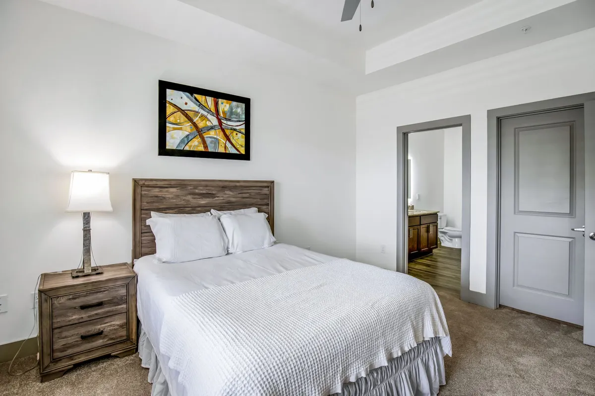 This image showcases a peaceful bedroom with a comfortable bed dressed in quality linens, a nightstand with a lamp, tasteful artwork above the bed, and a visible doorway leading to a bathroom. Natural light gently fills the room, enhancing the serene atmosphere.